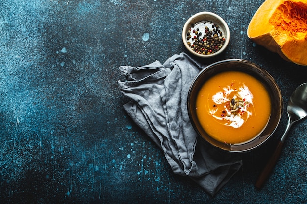 Sopa de creme de abóbora caseira servida em uma tigela de cerâmica marrom sobre fundo de pedra escura com colher decorada com abóbora fresca cortada, vista superior. Comida tradicional de outono, espaço para texto