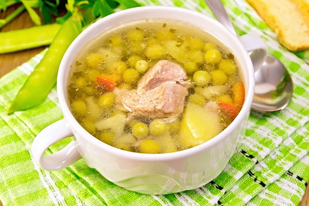 Sopa de ervilhas com carne e batatas em uma tigela branca, pão, colher, vagens em guardanapo verde no fundo das pranchas de madeira