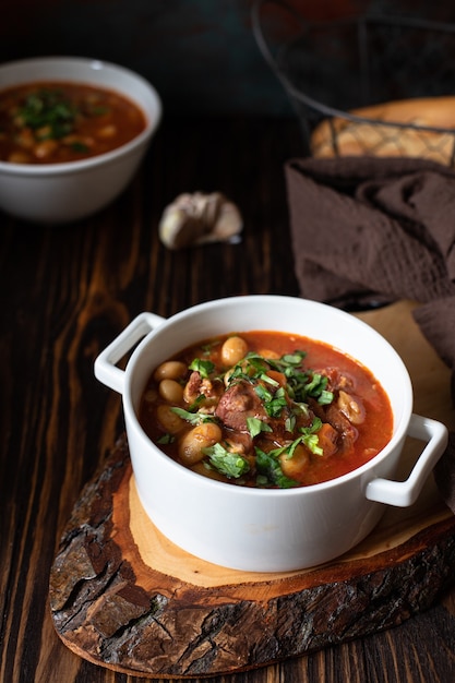 Sopa de feijão com carne e legumes servida em uma tábua rústica e mesa de madeira com pão e alho. Sopa tradicional dos Balcãs Pasulj (Grah). Close-up, foco seletivo