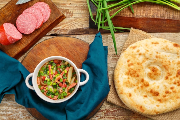 Sopa de feijão de cozinha judaica cholit em um guardanapo azul ao lado de salsicha de pão e cebolas verdes e uma colher de pau