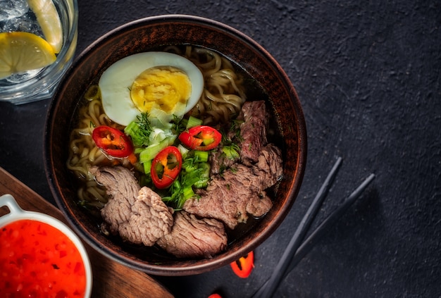 Sopa de macarrão asiático com carne, ovo, pimenta vermelha e ervas em uma mesa escura, vista superior