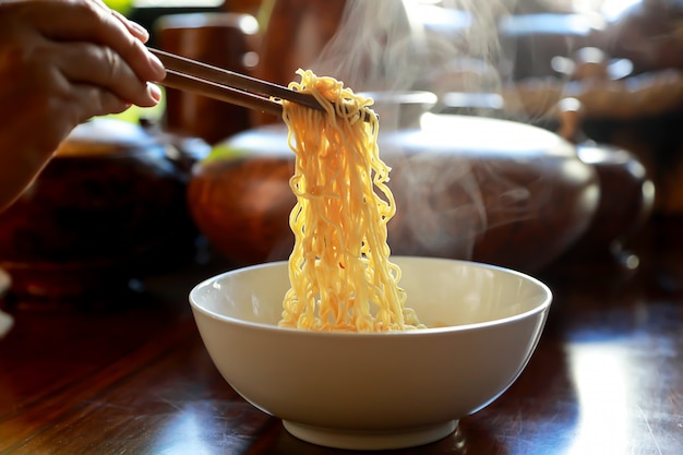 Sopa de macarrão instantâneo com pauzinhos na mão no fundo da mesa de madeira com fumaça.