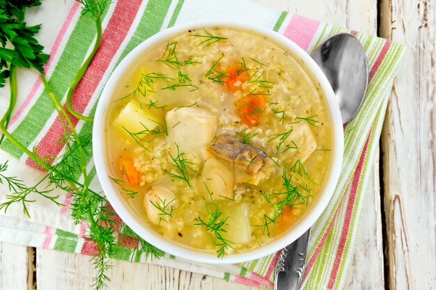 Sopa de peixe kulesh com painço, batata e cenoura em uma tigela branca em um guardanapo, salsa, endro em uma placa de madeira de fundo por cima