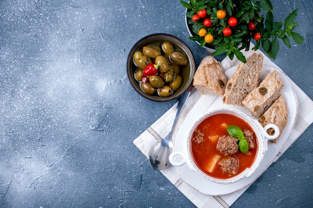 Sopa de tomate em um prato branco com bolas de carne, sobre um fundo de pedra com pão e azeitonas