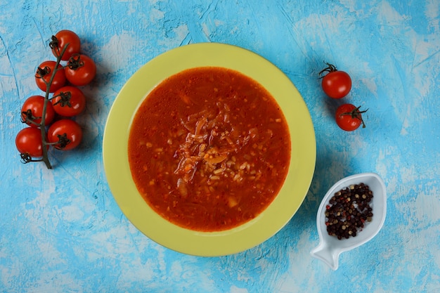 Sopa deliciosa, saborosa de tomate no prato verde sobre fundo azul. Tomate e prato decorativo com pimenta ao lado da refeição.