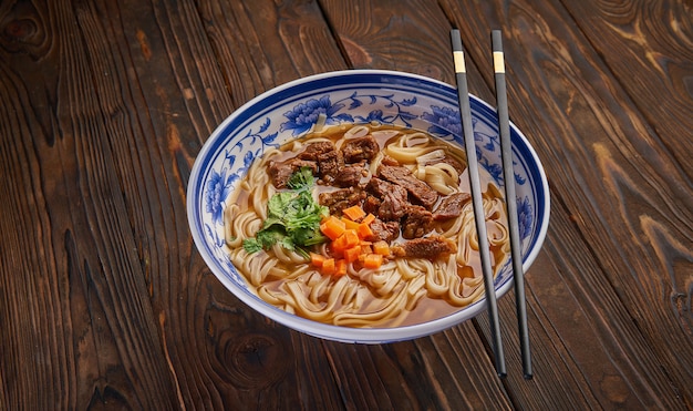 Sopa de fideos con carne china en tazón azul tradicional, hierbas frescas y zanahoria en rodajas, par de palillos en la mesa de madera oscura y copie el espacio. Vista superior del concepto de comida asiática