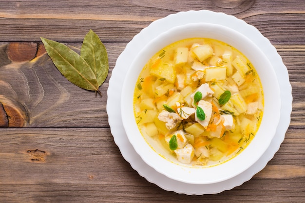 Sopa fresca de caldo de galinha com batatas e ervas em uma tigela branca e louro em uma mesa de madeira. Vista do topo