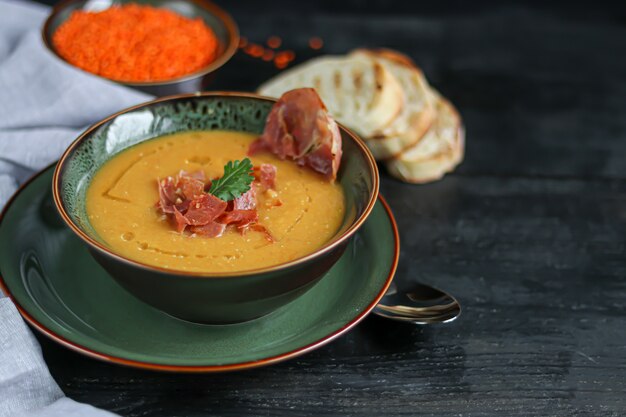 Sopa de lentejas sobre un fondo de madera. Servido con tocino en rodajas y hierbas. Cerca hay pedazos de chapata.