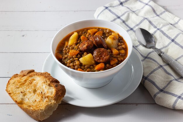 Sopa de lentejas tradicional con piernas, zanahorias y chorizo en un plato blanco sobre un fondo blanco