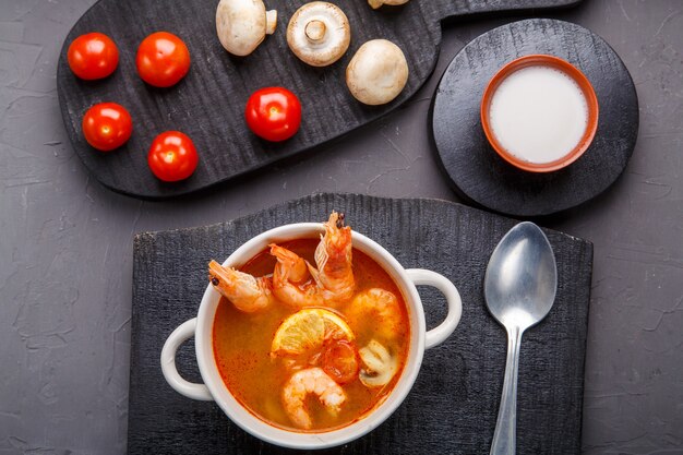 Sopa de ñame de tom con camarones en una sopera sobre un fondo de hormigón junto a un cuenco con tomates de leche de coco y champiñones sobre un soporte de madera.
