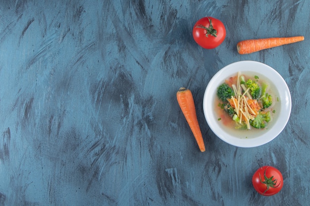 Sopa de pollo con brócoli y zanahoria en un bol junto a verduras, sobre fondo azul.