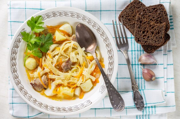Sopa de pollo casera con fideos y verduras con hojas de apio en una servilleta de algodón, con una cuchara y un tenedor. Pedazos de pan de centeno en una servilleta.