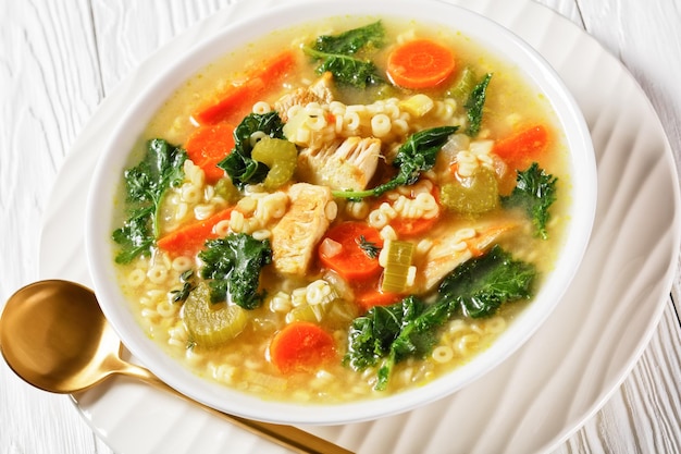 Sopa de pollo con col rizada y pasta anellini y verduras en un recipiente blanco con cuchara dorada sobre una mesa de madera, cocina italiana