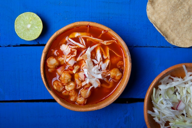 Sopa de pozole rojo tradicional mexicana con tostada y limones sobre un fondo azul.