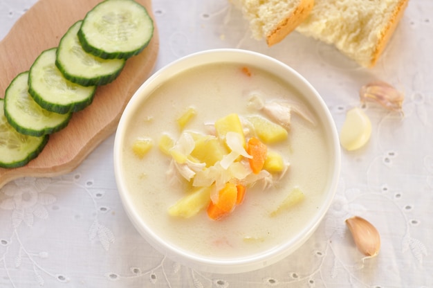 Sopa de queso con pollo y verduras en un tazón blanco, pepino y ajo en una servilleta textil. Vista superior