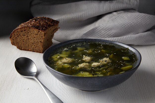 Sopa rústica de abobrinha com almôndegas com um pedaço de pão preto com sementes de cominho em uma mesa branca contra o fundo de um pano de prato cinza na luz da manhã