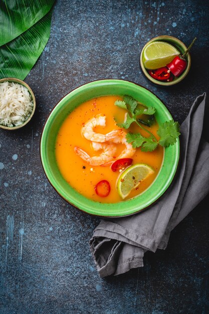 Sopa tailandesa saudável tradicional tom yum com camarão, limão, coentro em uma tigela sobre fundo rústico com arroz branco, tiro aéreo. Conceito de comida tailandesa autêntica com espaço de cópia