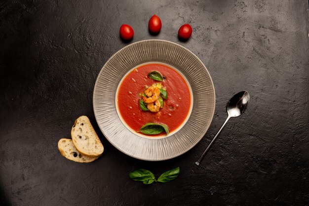 Sopa de tomate clásica española con gambas, hojas de albahaca, tomates cherry y trozos de ciabatta sobre una mesa de cocina de piedra negra