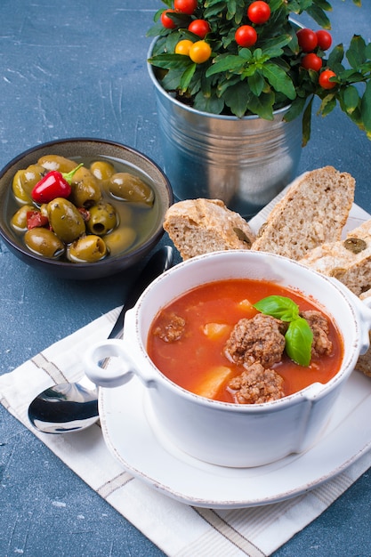 Foto sopa de tomate en un plato blanco con albóndigas, sobre un fondo de piedra con pan y aceitunas