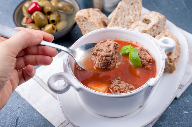Sopa de tomate en un plato blanco con albóndigas, sobre un fondo de piedra con pan y aceitunas