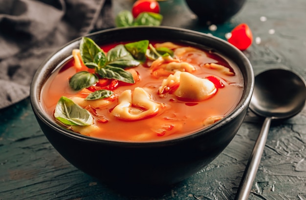 Sopa de tomate con tortellini en un tazón negro sobre fondo oscuro.