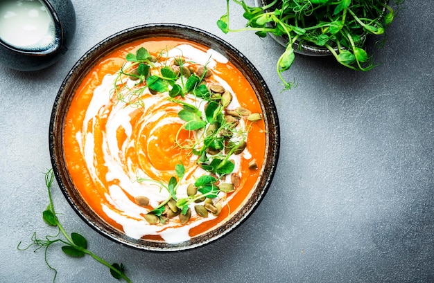 Sopa vegana de calabaza y zanahoria con semillas de calabaza, crema de coco y brotes de guisantes Comida lenta vegetariana saludable de invierno u otoño Tazón de sopa sobre fondo de mesa gris Vista superior