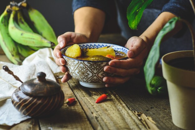 Sopa de verduras latinoamericana, manos agarrando el plato. Ingredientes zanahoria, chayote, boniato, pimiento plátano verde.