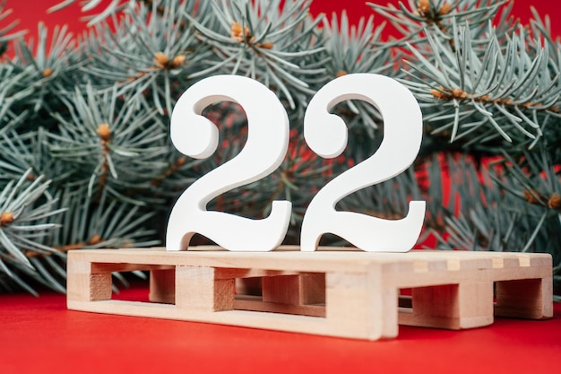 Soporte de números 22 de madera blanca con exuberantes ramas de árboles de Navidad verdes en palet sobre fondo rojo. Feliz Navidad 2022