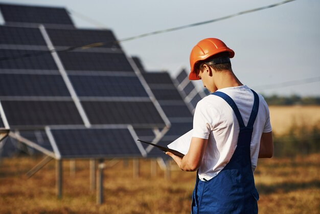 Soportes con bloc de notas. Trabajador de sexo masculino en uniforme azul al aire libre con baterías solares en un día soleado.