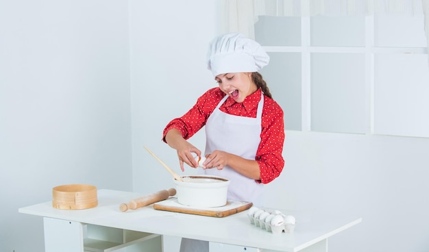 Sorgfältige Herangehensweise glückliches Kind, das in der Küche kocht, backt Kekse in der Küche, professionelles und erfahrenes Bäckerkind in Kochuniform und Hut, Teenager-Mädchen bereitet Teig zu und macht Kuchen nach Rezeptzeit zum Essen