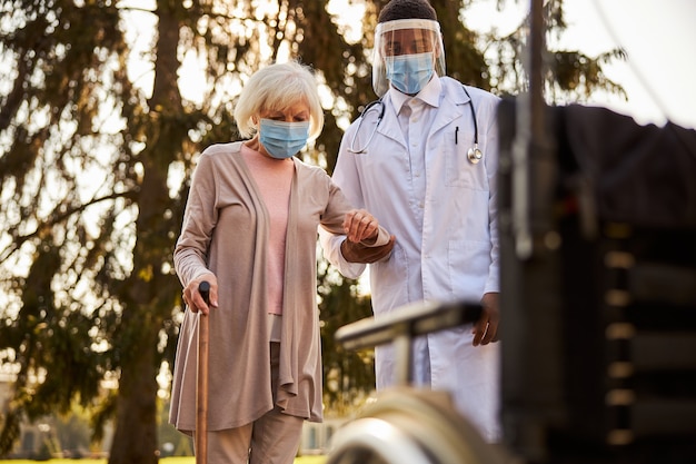 Sorgfältige medizinische Person mit Gesichtsschutz und Maske, die eine ältere Frau auf dem Weg zum Rollstuhl unterstützt