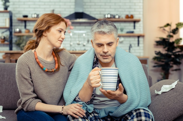 Sorgfalt und Liebe. Schöne nette Frau, die ihren Mann ansieht, während sie ihn während seiner Krankheit unterstützt supporting