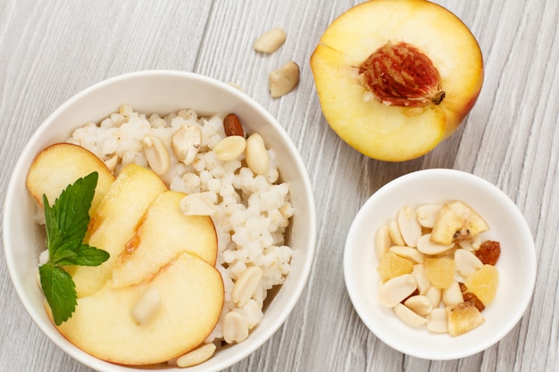 Sorghumbrei mit Pfirsichstücken, Cashewnüssen und Mandeln in Porzellanschalen, ein frischer Pfirsich auf den grauen Holzbrettern. Veganer glutenfreier Sorghumsalat mit Früchten.