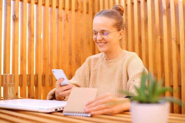 Sorglose Frau in beigem Pullover sitzt in einem Straßencafé und arbeitet am Laptop mit Telefon und Papierhobel, plant ihre Arbeit und genießt die freiberufliche Arbeit