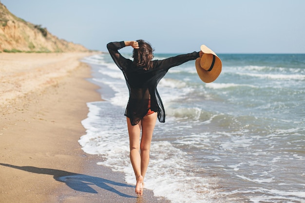 Sorglose Frau mit Hut, die am Sandstrand bei Meereswellen spazieren geht und Sommerferien entspannt