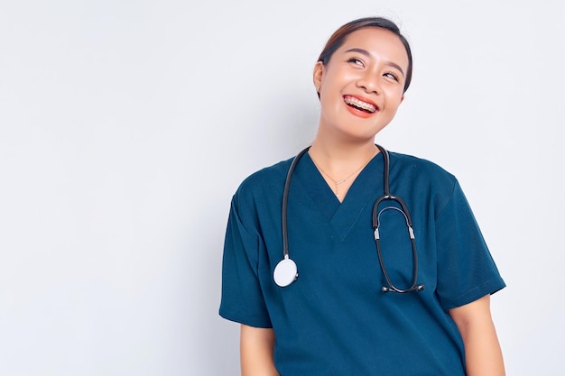 Sorglose junge asiatische Frau Krankenschwester in blauer Uniform mit einem Stethoskop-Praktikanten, der Spaß hat, glückliches Mittagspause-Kichern zu lachen, das auf weißem Hintergrund isoliert wird. Healthcare-Medizin-Konzept