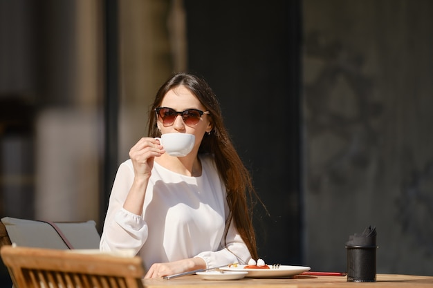 Sorglose junge Frau, die auf der Terrasse des Cafés sitzt und Cappuccino trinkt