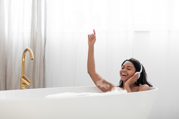 Sorglose junge schwarze Frau, die Musik hört, während sie mit Schaum badet