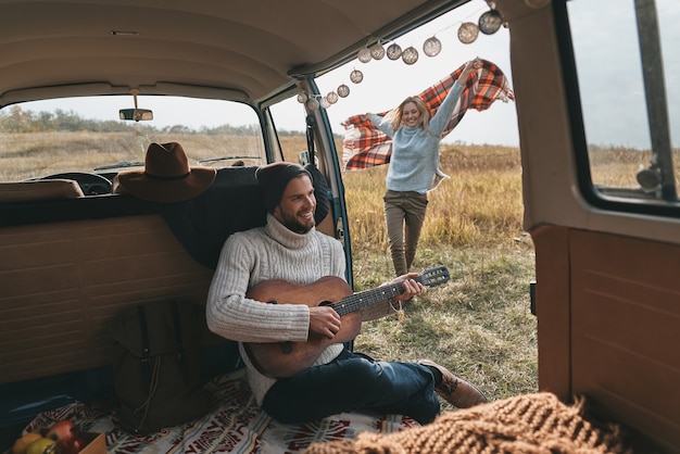 Sorglose Reise. Hübscher junger Mann, der Gitarre für seine schöne Freundin spielt, während im Retro-Art-Minivan sitzt