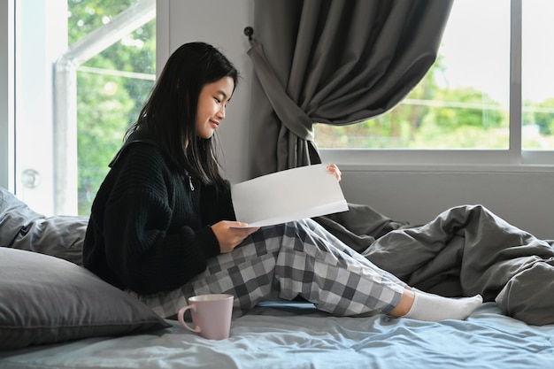 Sorgloser Asiate sitzt in einem gemütlichen Schlafzimmer und liest ein Buch. Freizeit- und Menschenkonzept