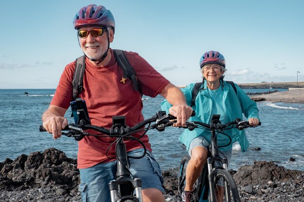 Sorgloses kaukasisches Seniorenpaar mit Helmen, die mit Elektrofahrrädern am Strand fahren Authentisches Lebenskonzept für ältere Menschen im Ruhestand Horizont über dem Meer