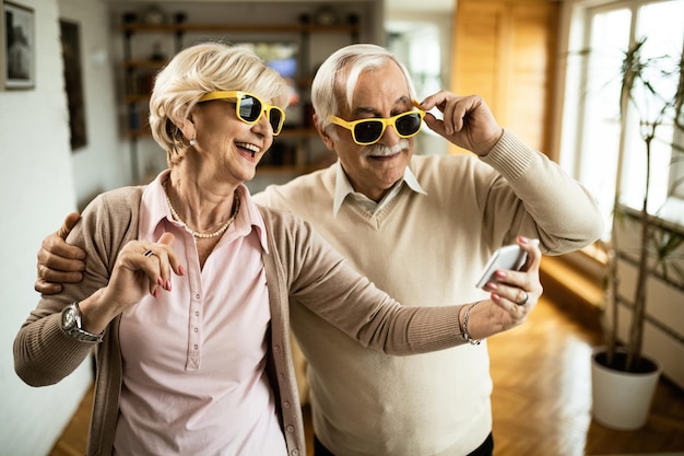 Sorgloses Seniorenpaar macht Selfie mit Smartphone, während es zu Hause eine Sonnenbrille trägt