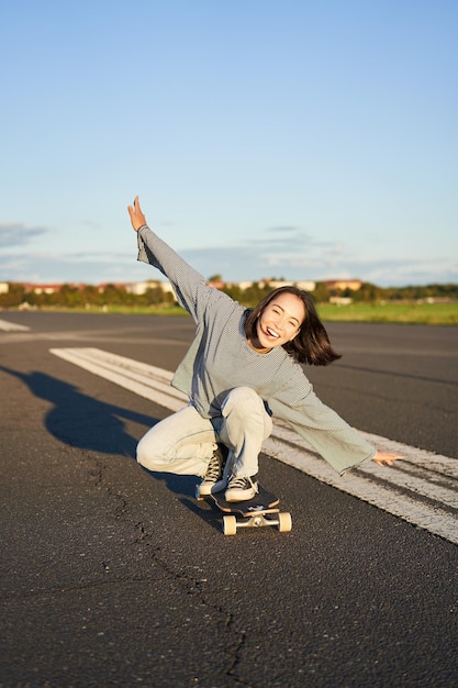 Sorgloses Skatermädchen auf ihrem Skateboard, das Longboard auf einer leeren Straße fährt, die Hände seitlich hält und