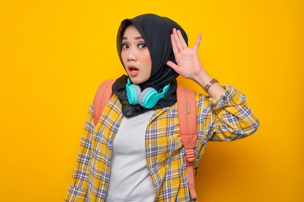 Sorprendente joven estudiante asiática musulmana con camisa a cuadros con auriculares y mochila cogidos de la mano cerca de sus oídos escuchando chismes sobre fondo amarillo Concepto de colegio universitario de la escuela de educación