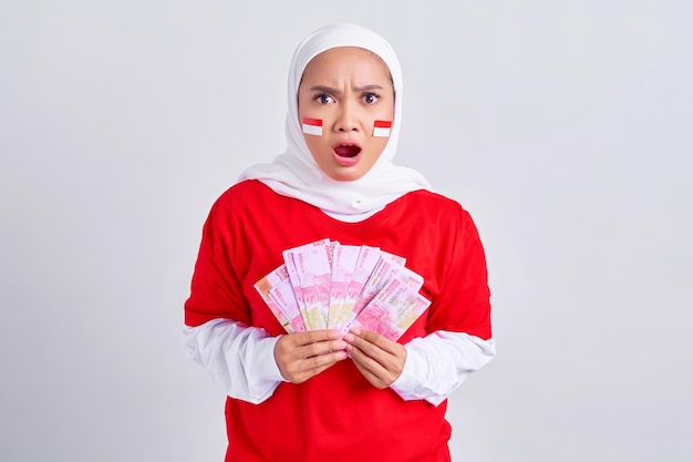 Sorprendente joven mujer musulmana asiática en camiseta blanca roja con dinero en efectivo en billetes de rupias indonesias aislado sobre fondo blanco Día de la independencia de Indonesia el 17 de agosto concepto