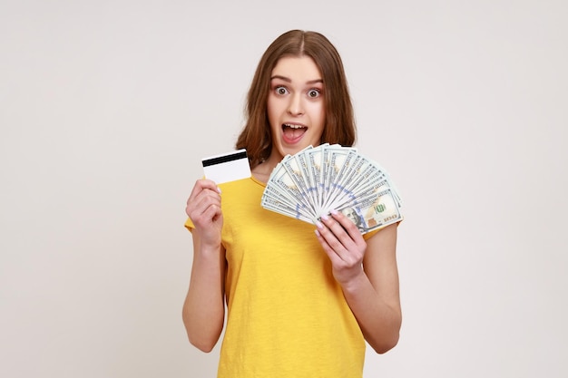 Sorprendida adolescente emocionada con cabello castaño en camiseta amarilla sosteniendo y mostrando billetes de dólar y tarjeta de crédito, sorprendida con el interés del depósito. Disparo de estudio interior aislado sobre fondo gris.