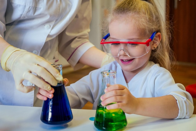 Sorprendida y asombrada, inteligente rubia colegiala química con anteojos y bata médica blanca haciendo experimentos científicos química mezclando diferentes soluciones químicas en el aula de laboratorio