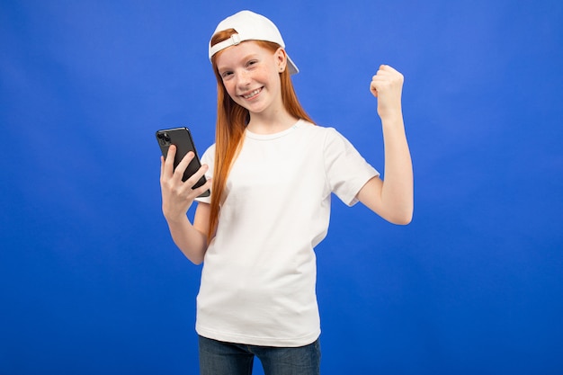 Sorprendida chica adolescente pelirroja en una camiseta blanca con un gadget para comunicarse en manos azules