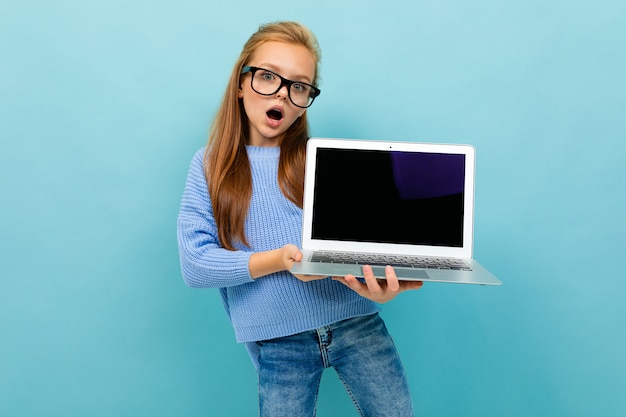 Sorprendida colegiala sorprendida sobre un fondo azul sostiene la computadora portátil con la pantalla hacia adelante