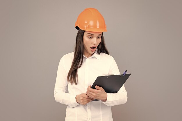 Sorprendida, conmocionada y emocionada mujer de negocios con clipboard en fondo gris Retrato de una trabajadora de la construcción con casco